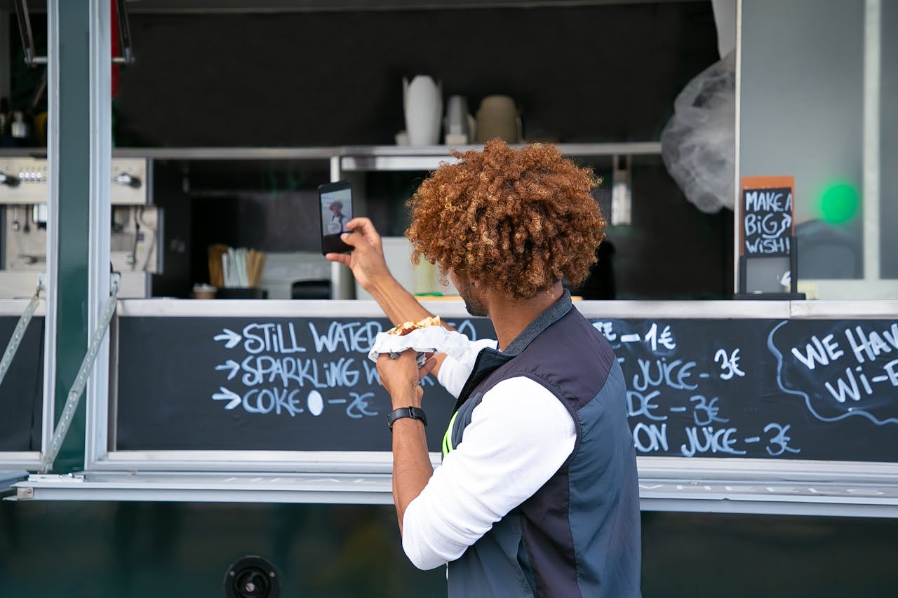 Side view unrecognizable African American male in casual clothes eating yummy sandwich near food truck and taking selfie on mobile phone